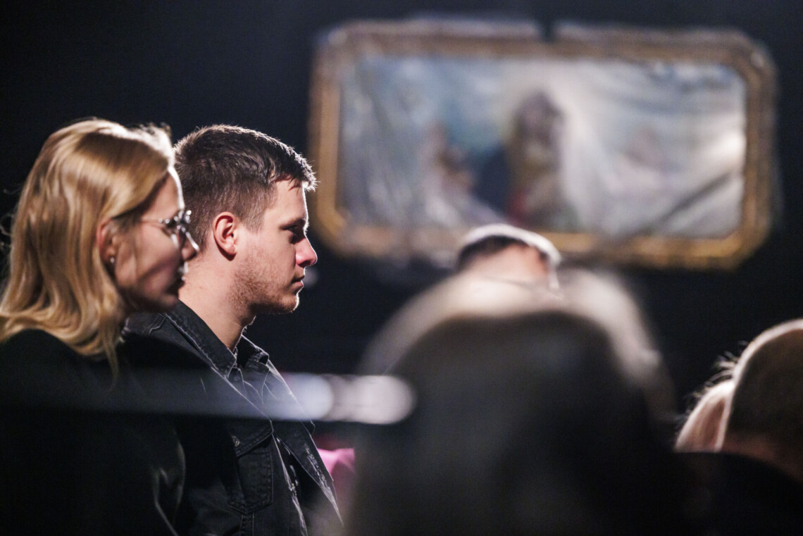 Osoby siedzące na widowni, ujęcie z boku, w tle rozmazany kontur dużego, prostokątnego obrazu