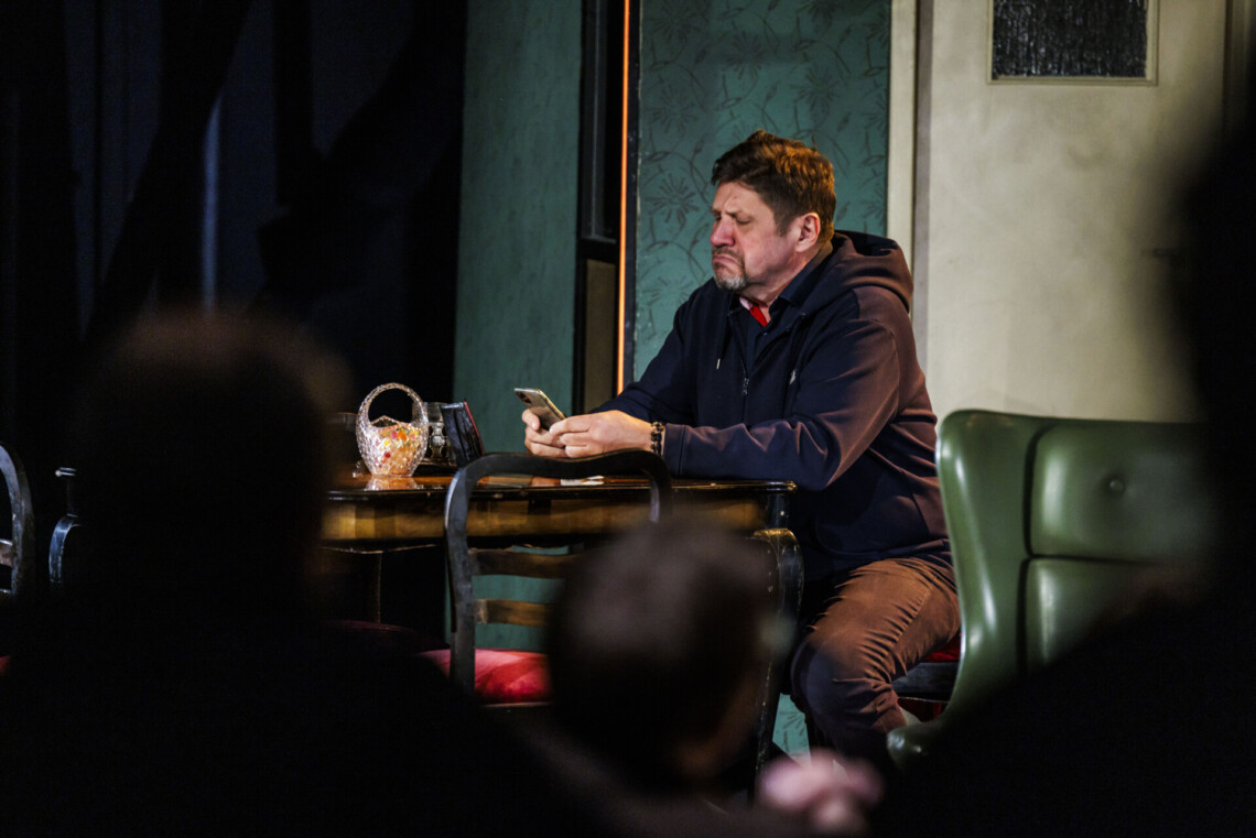 Aktor na scenie w trakcie spektaklu - mężczyzna siedzi przy stole, przegląda telefon