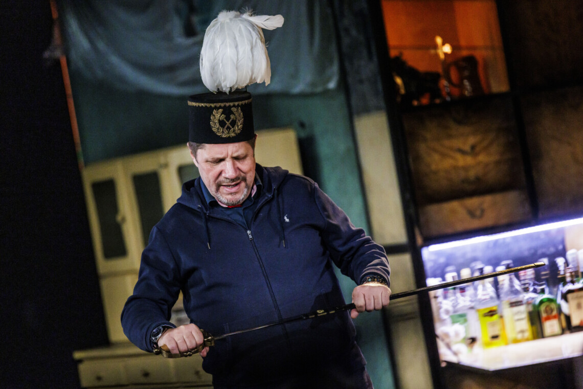 Mężczyzna w górniczej czapce z białym pióropuszem, w dłoniach trzyma szablę, którą wysuwa z pochwy, wzrok ma skierowany w dół, na szablę