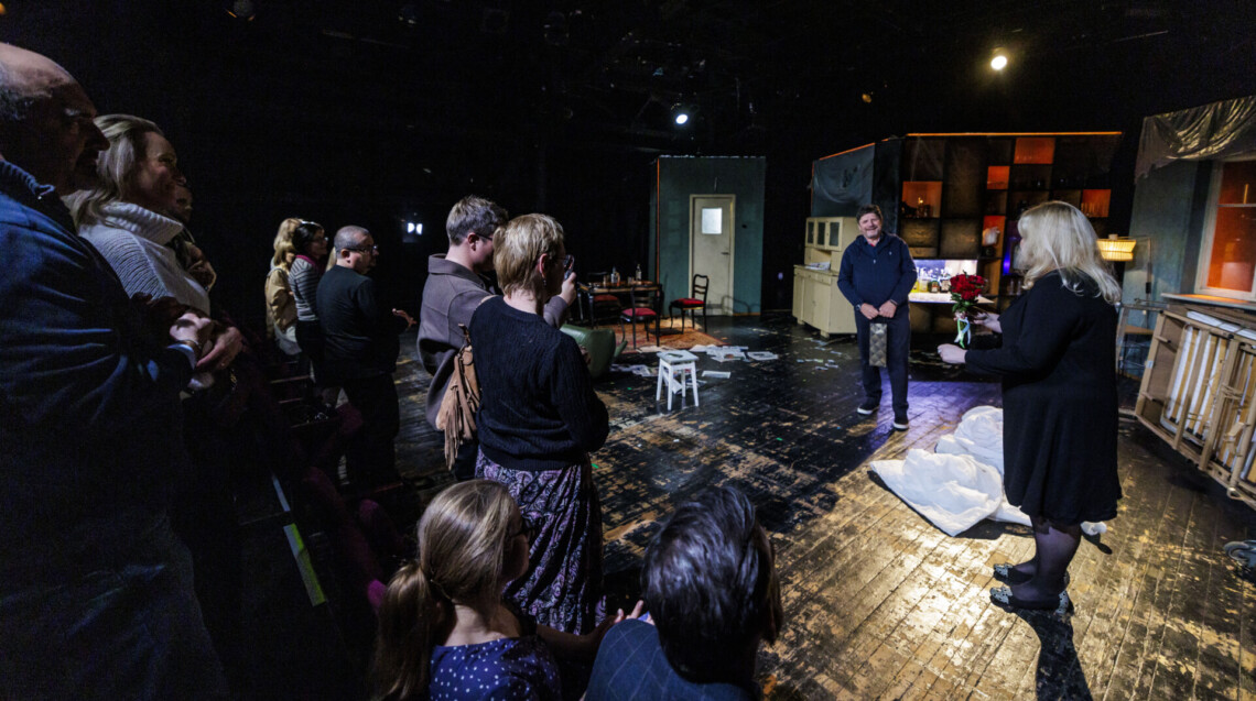 Na scenie aktor trzymający w dłoniach torebkę prezentową, pochodzi do niego kobieta z czerwonymi kwiatami, z lewej strony osoby stoją na widowni; ujęcie pod kątem