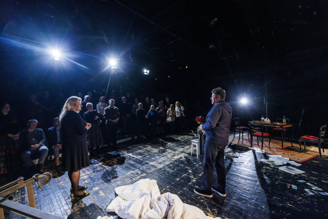 Aktor trzyma w dłoniach kwiaty, z lewej stronie na scenie stoi kobieta, przed nimi osoby stojące na widowni; ujęcie od tyłu