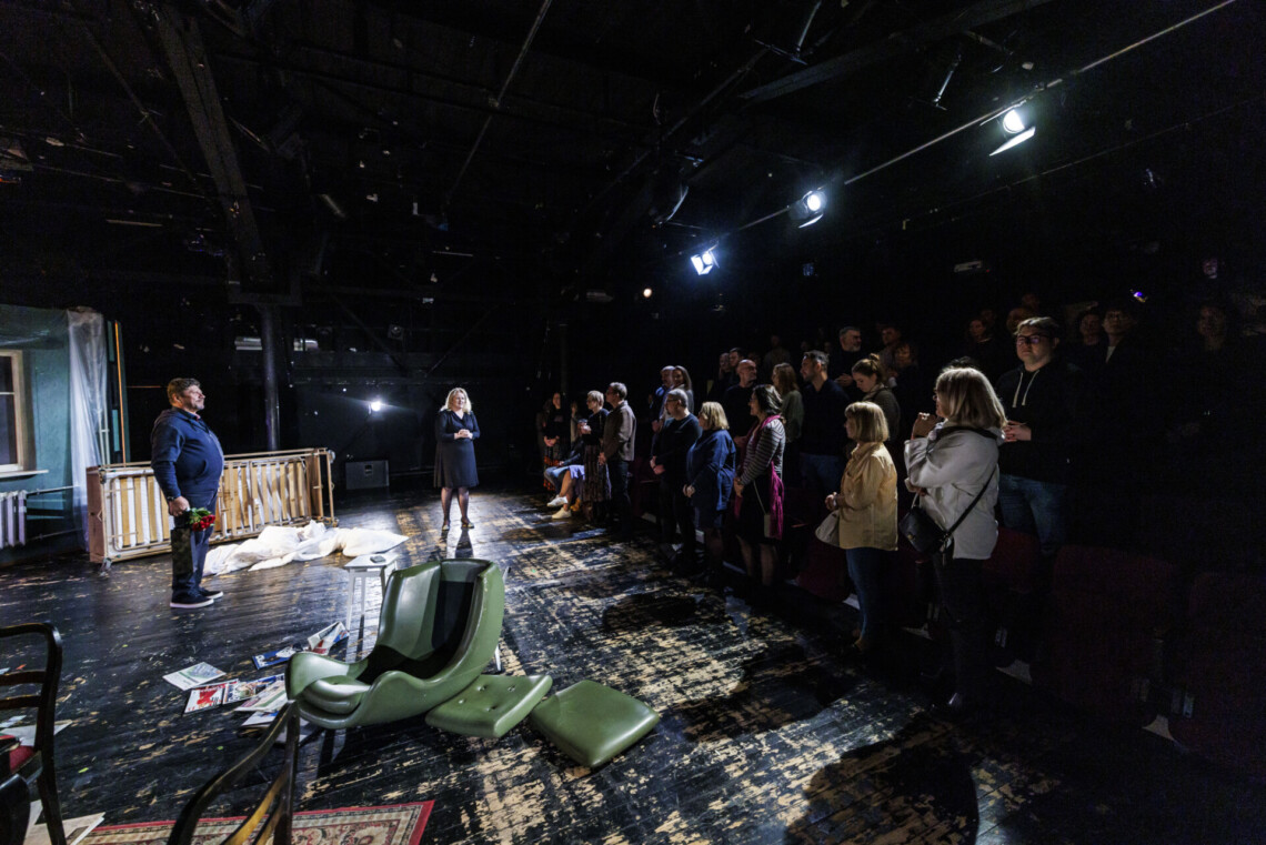 Na cenie w centrum stoi aktor, w oddali kobieta ubrana na czarno, z prawej strony osoby stoją na widowni, ujęcie z boku