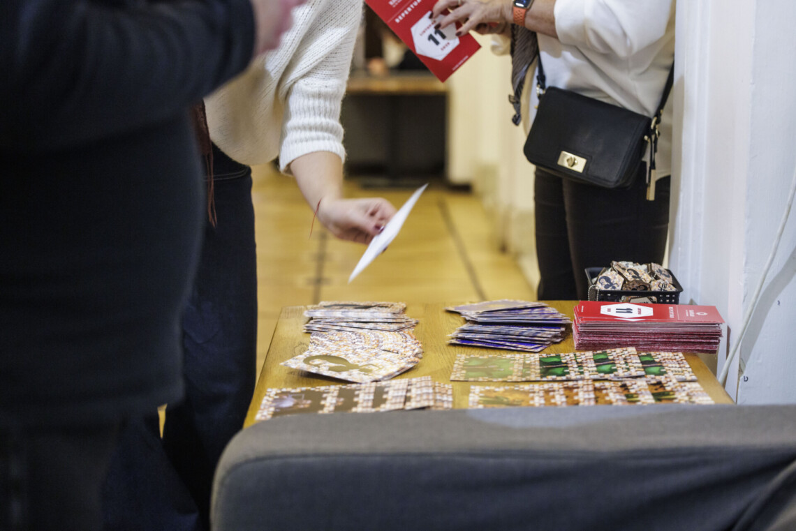 Na stole leżą pocztówki promocyjne oraz ulotka repertuarowa Teatru Śląskiego, dwie osoby pochylają się nad stolikach - widoczne są ich postury od ramion w dół