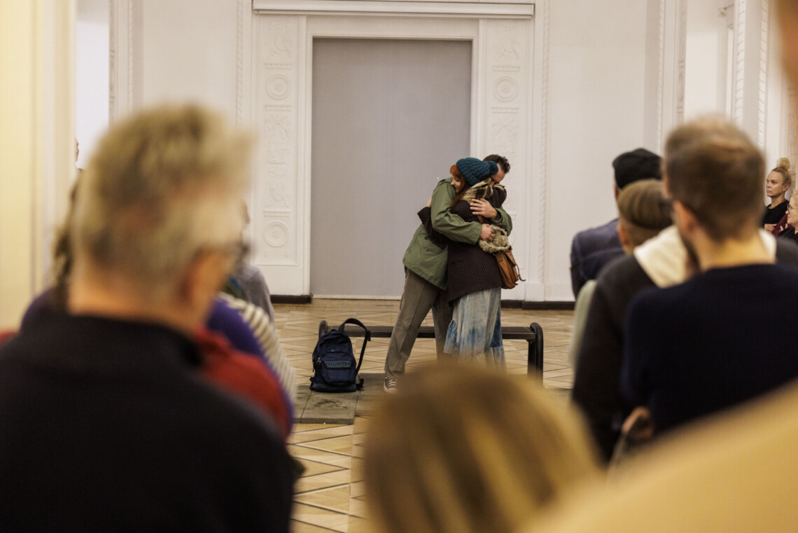 Kobieta i mężczyzna obejmują się, stoją przy ławce, scena ze spektaklu, na pierwszym planie rozmazane postacie widzów od tyłu