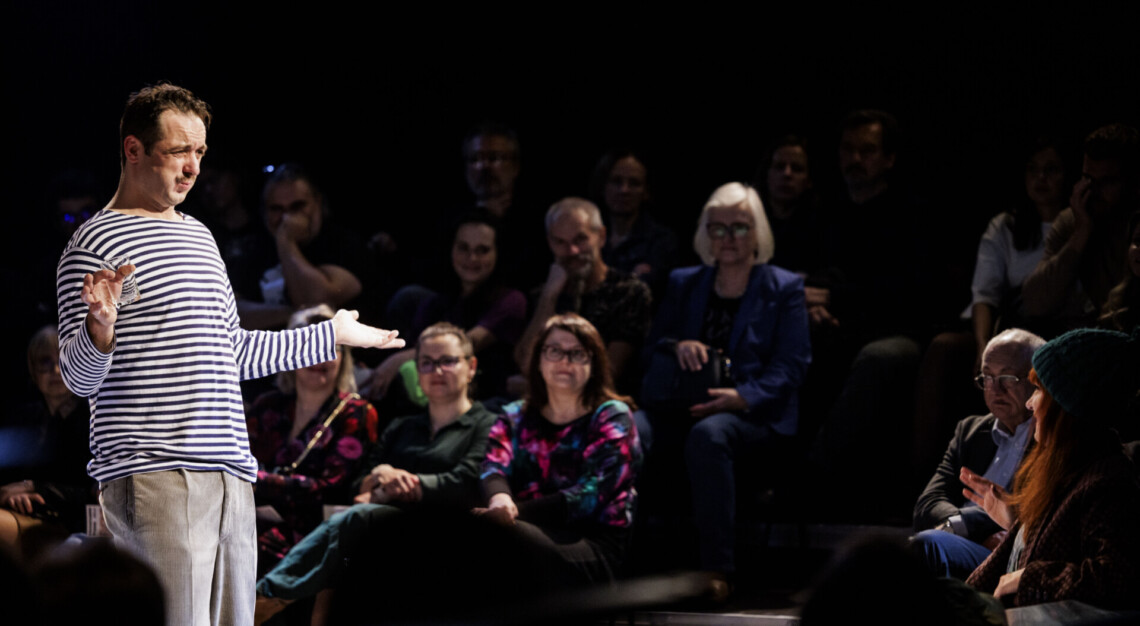 Mężczyzna w koszulce w biało-grantowe paski stoi z rozłożonymi na bok rękami, w prawej dłoni trzyma szklankę, na przeciwko niego siedzi kobieta, na drugim planie osoby siedzące na widowni