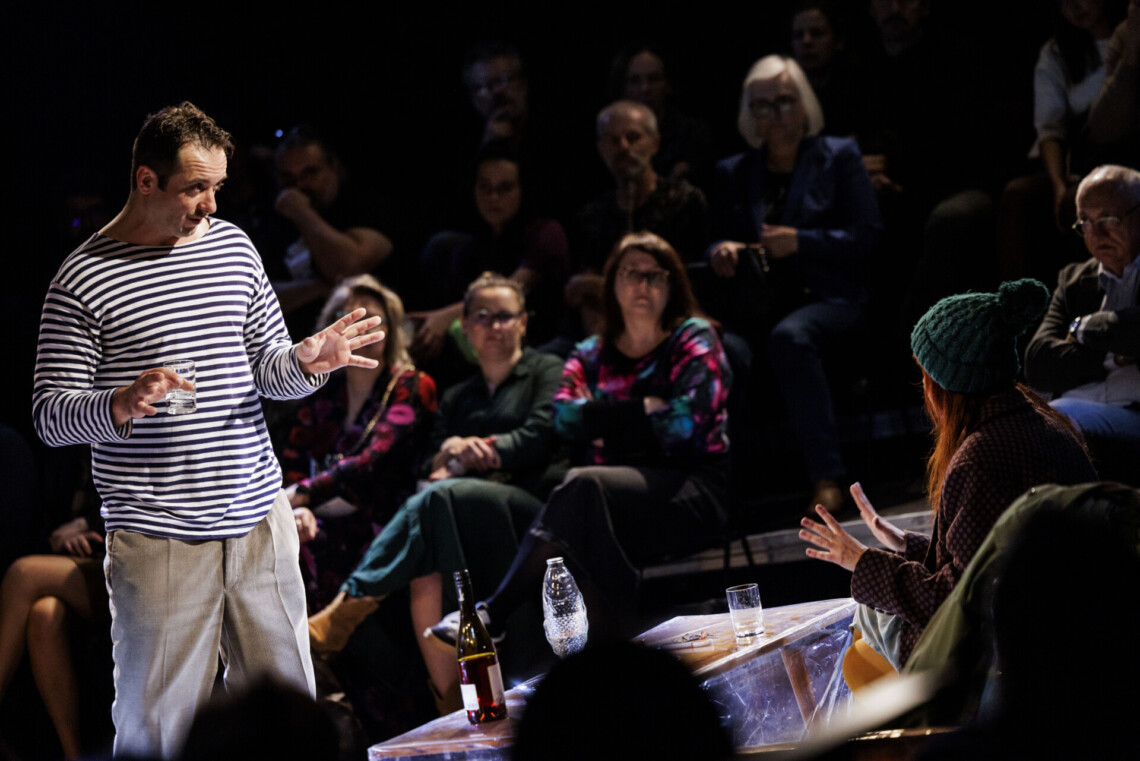 Przed stolikiem stoi mężczyzna ze szklanką w dłoni, na kanapie za stolikiem siedzi kobieta, znajdują się oni naprzeciwko siebie