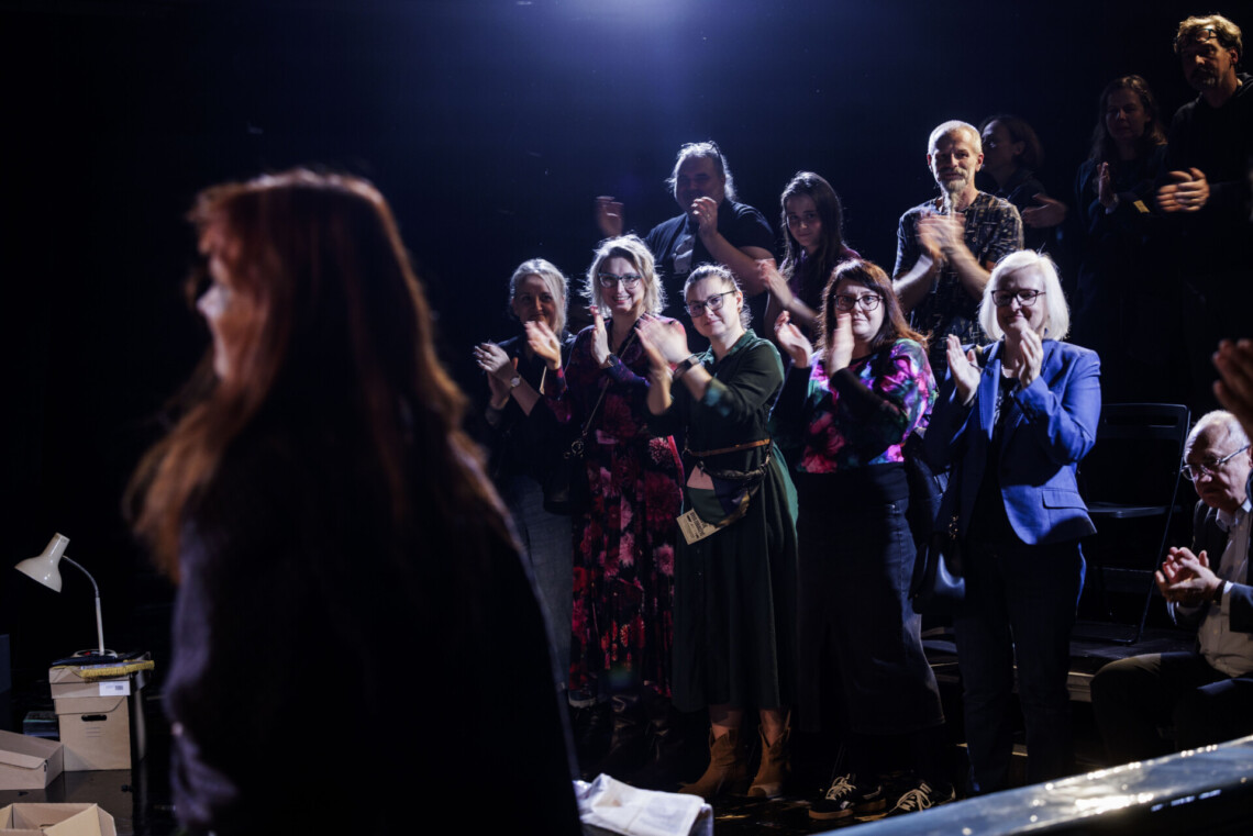 Na pierwszym planie niewyraźna postać aktorki od tyłu, na drugim planie osoby stojące na widowni w trakcie oklasków