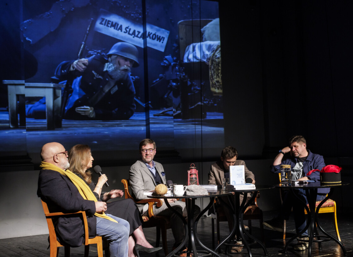 Pięć osób siedzących za krzesłach przy trzech okrągłych stolikach, za nimi na ekranie wyświetlony kadr - mężczyzna w hełmie leżący na brzuchu na podłodze, za nim napis "Ziemia Ślązaków!"