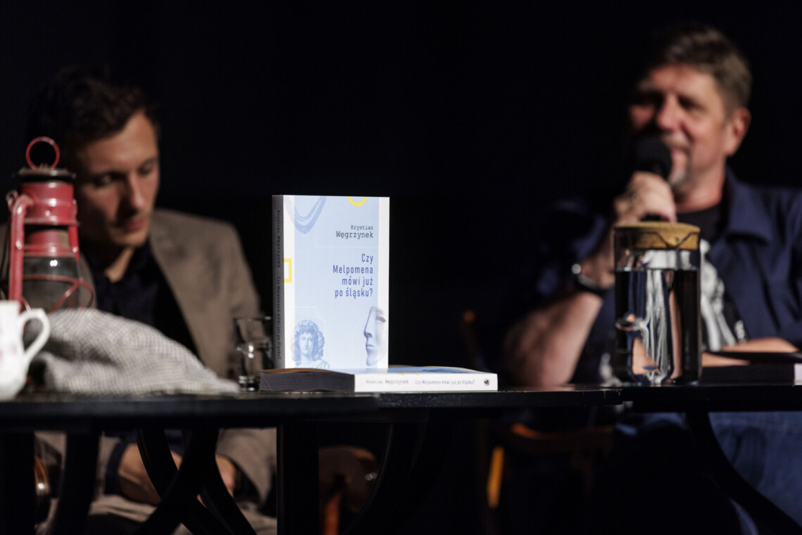 Na stoliku pionowo stoi książka "Czy Melpomena godo po śląsku", w tle rozmazane postacie dwóch osób