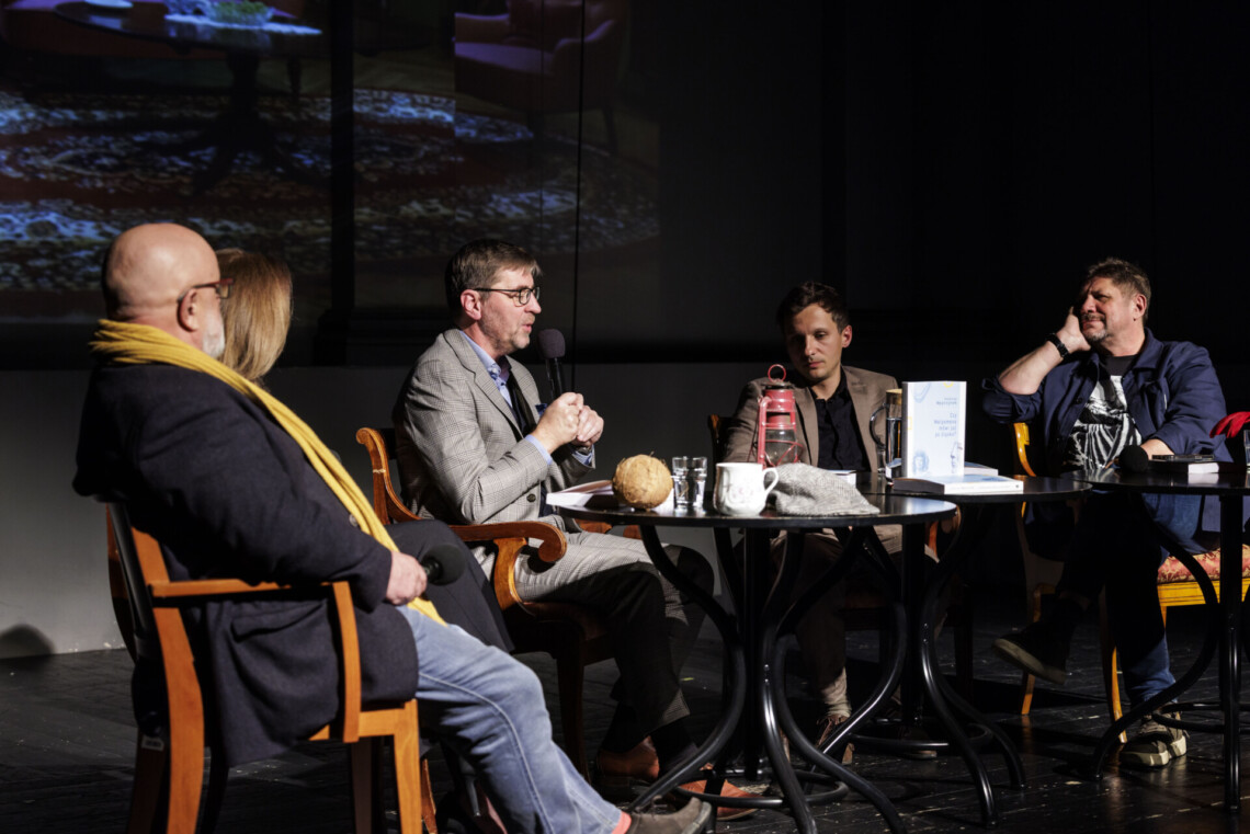 Pięć osób siedzących za krzesłach przy trzech okrągłych stolikach, do mikrofonu przemawia Krystian Węgrzynek siedzący pośrodku