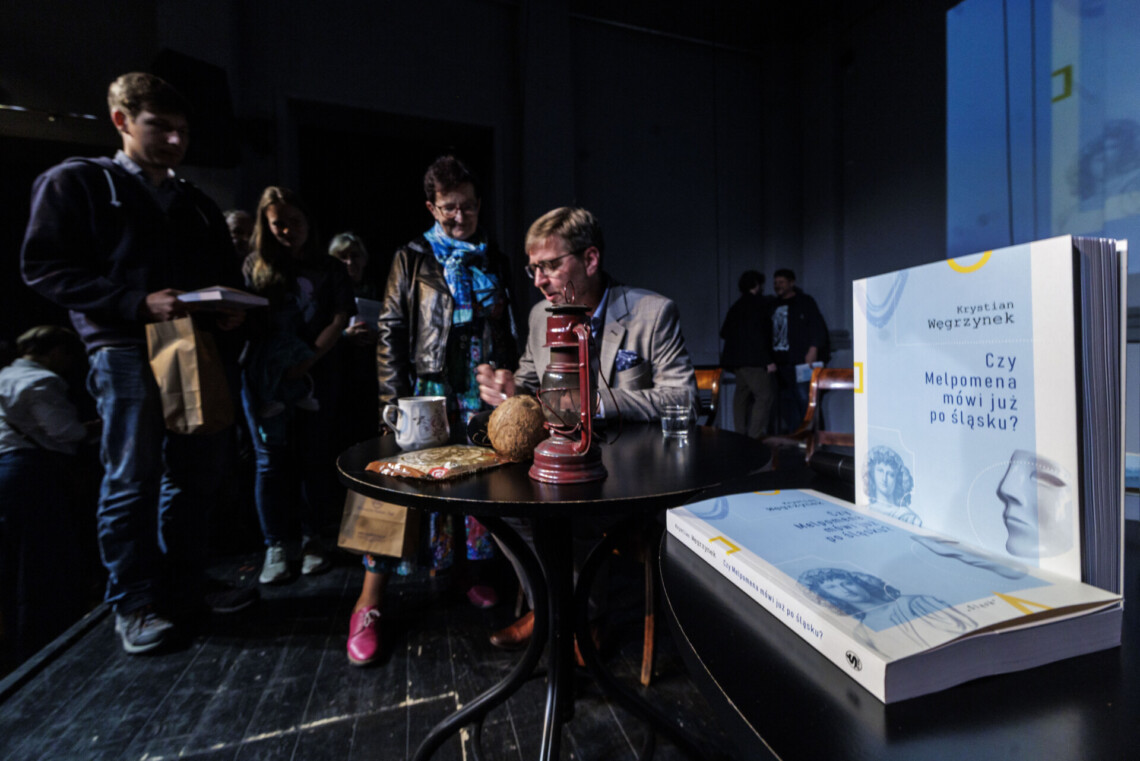Na pierwszym planie dwa egzemplarze książki "Czy Melpomena godo po śląsku" - jedna leży druga stoi, okładka jest jasnoniebieska, na drugim planie Krystian Węgrzynek podpisujący swoją książkę