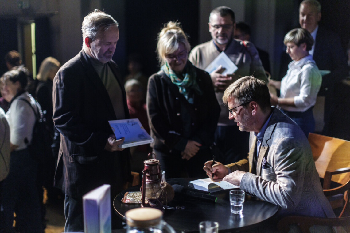 Krystian Węgrzynek podpisujący swoją książkę, obok stoją kobieta i mężczyzna