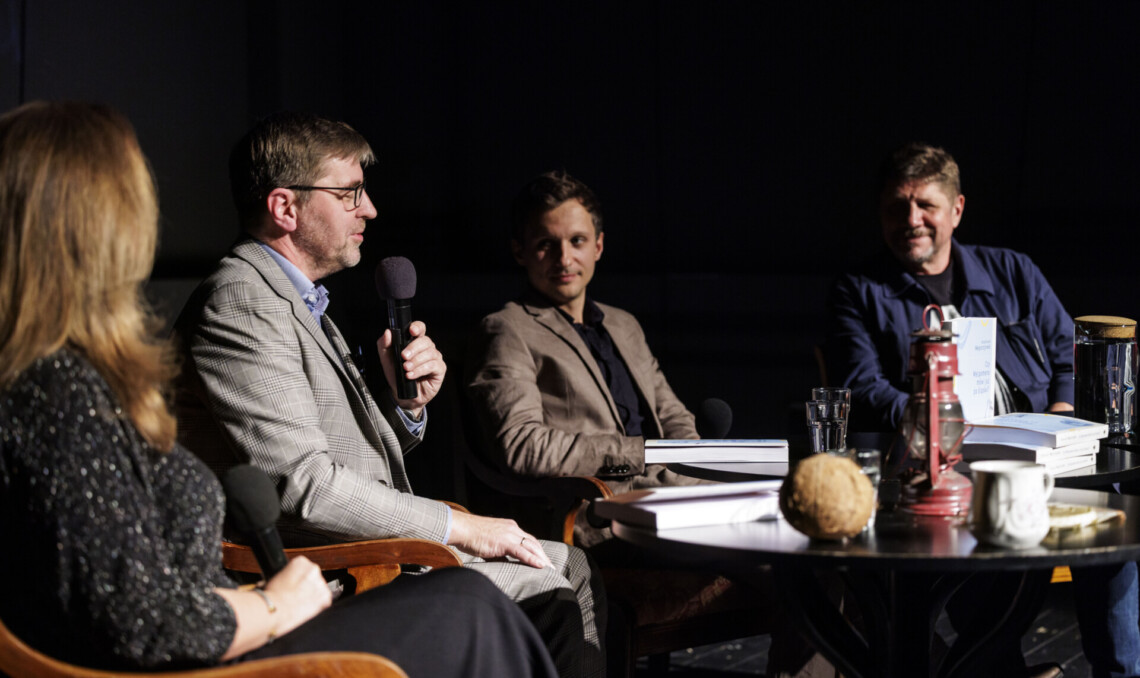 Cztery osoby siedzące za krzesłach, do mikrofonu przemawia Krystian Węgrzynek; przybliżenie