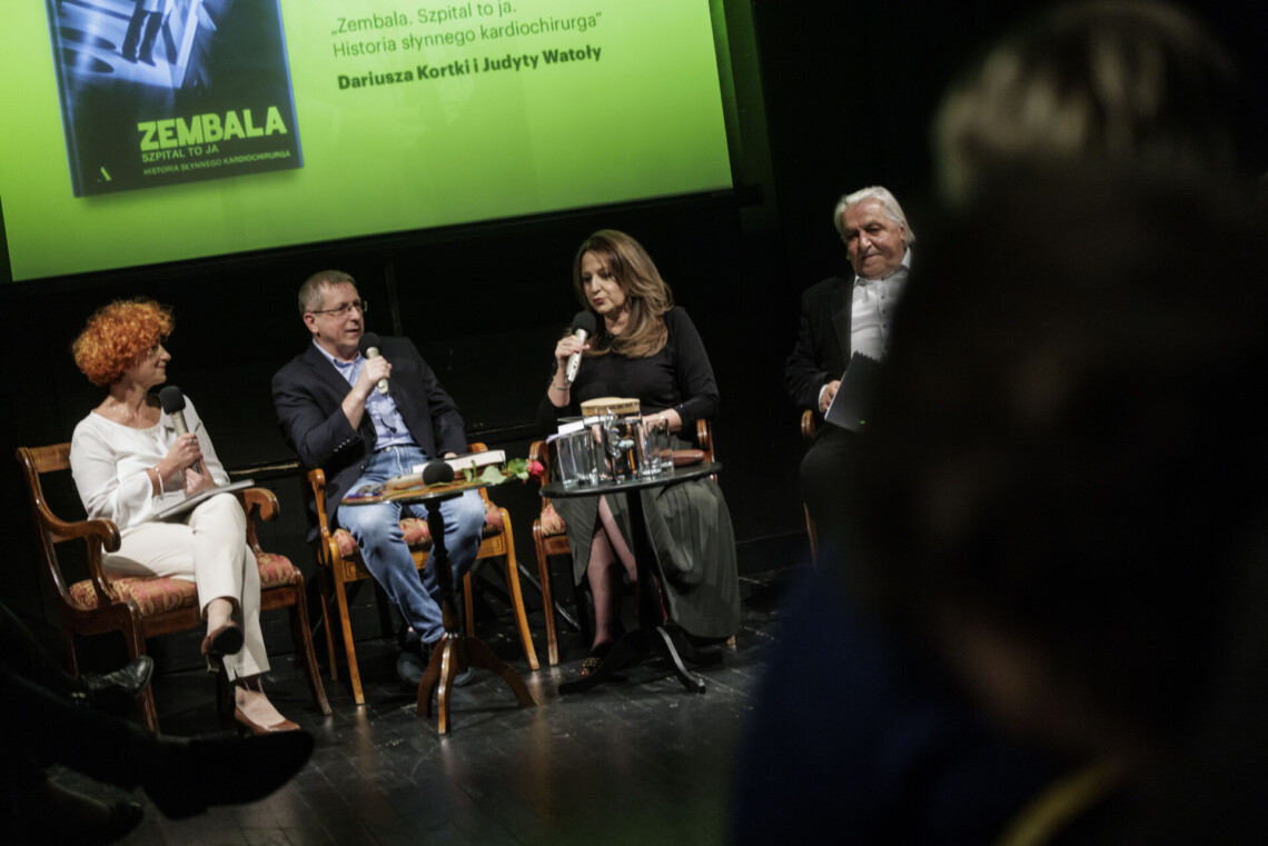 Cztery osoby siedzące na krzesłach na scenie, trzy z nich trzymają mikrofony, za nimi fragment żółtego plakatu promującego spotkanie