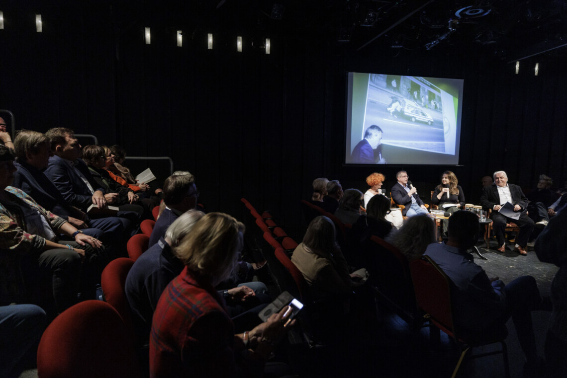 Z lewej osoby siedzące na widowni, z prawej osoby na scenie, za nim na ekranie zdjęcie mężczyzny z mikrofonem stojącego bokiem przed dużym zdjęciem karetki starego typu na ulicy