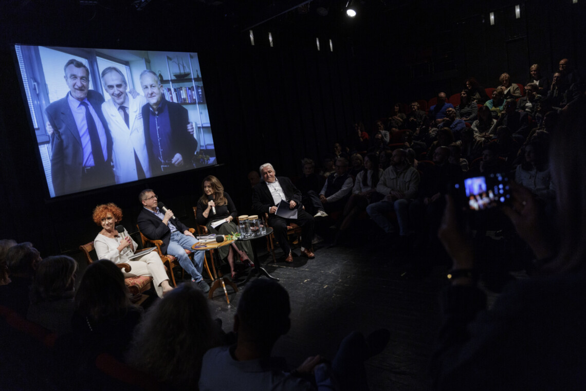 W centrum cztery osoby siedzące na widowni, za nimi na ekranie trzech mężczyzn pozujących do zdjęcia, wokół sceny pogrążona w ciemności widowni