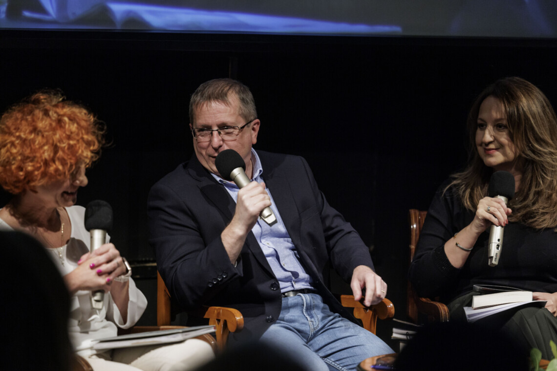 Dariusz Kortko przemawia do mikrofonu, po jego bokach siedzą kobiety