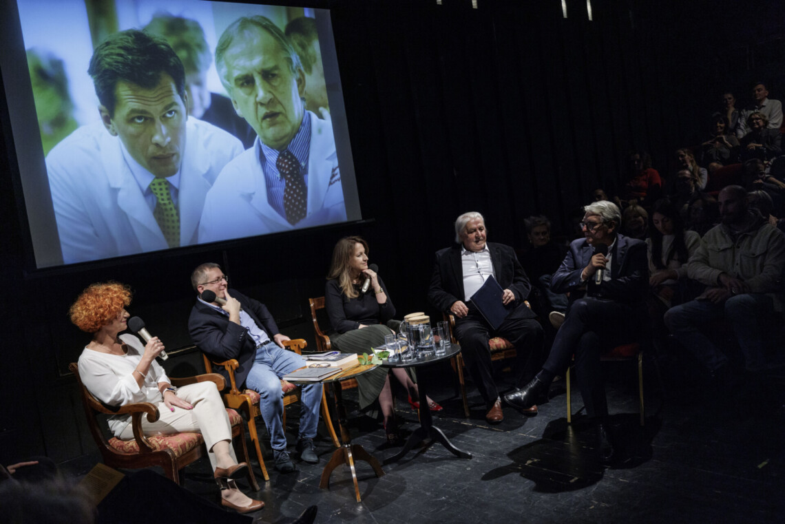 Pięć osób siedzących na scenie na krzesłach za dwoma okrągłymi stolikami, za nimi na ekranie dwóch mężczyzn w koszulach i krawatach, na której mają biały fartuch lekarski, jednym z nich jest profesor Zembala