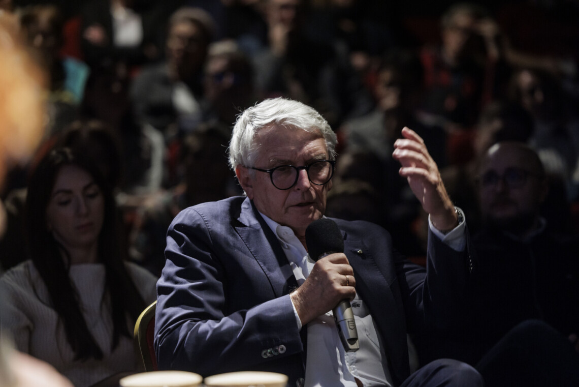 Starszy mężczyzna siedzący na krześle przemawia do mikrofonu, lewą rękę ma uniesioną w górę