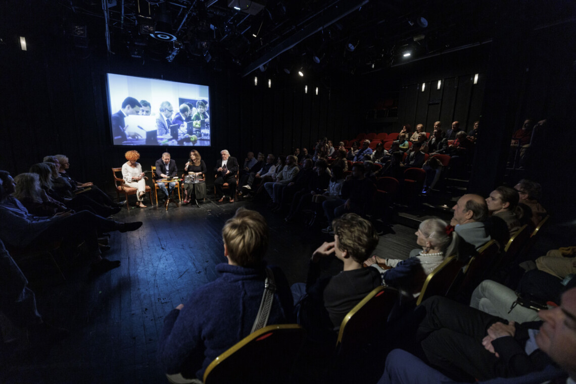 W centrum scena, na której na krzesłach siedzą cztery osoby, za nimi ekran z wyświetlonym zdjęciem osób z laptopami przy długim stole, z prawej i lewej strony sceny osoby siedzące na widowni, również na wprost osób na scenie siedzą widzowie na dostawionych krzesłach