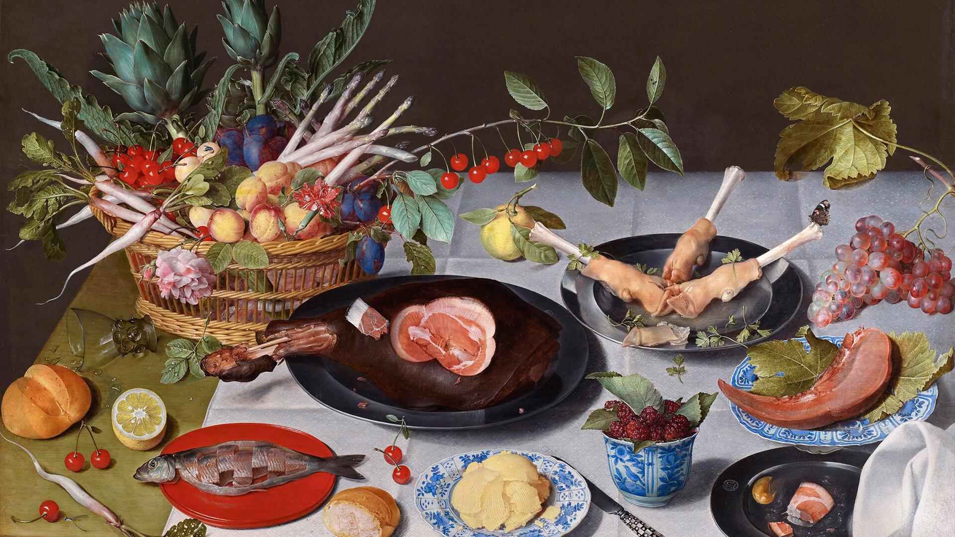 Obraz Jacoba van Hulsdoncka - stół przykryty białym obrusem, na nim z lewej u góry duży kosz z warzywami i owocami – szparagi, karczochy, śliwki, brzoskwinie, czereśnie, liście, poniżej na stole cytryna, bułka i ryba na czerwonym talerzu, w centrum duży półmisek z szynką, z prawej czarny talerz ze świńskimi nóżkami, niebiesko-biały z masłem lub serem, porcelanowy kubek z jeżynami, po prawej winogrona i plaster mięsa lub arbuza na liściu, tło ciemne, kompozycja bogata i szczegółowa, obraz realistyczny