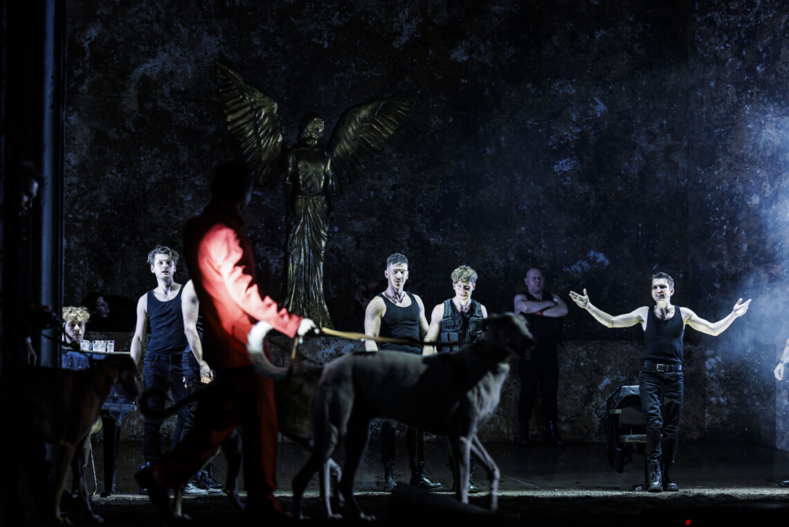 Na pierwszym planie przechadza się mężczyzna w czerwonym garniturze z dwoma psami na smyczy, na drugim planie stoją mężczyźni ubrani na czarno