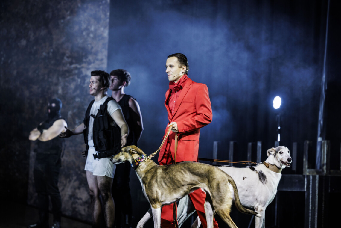 Mężczyzna w czerwonym garniturze z dwoma psami na smyczy, na drugim planie stoi trzech mężczyzn