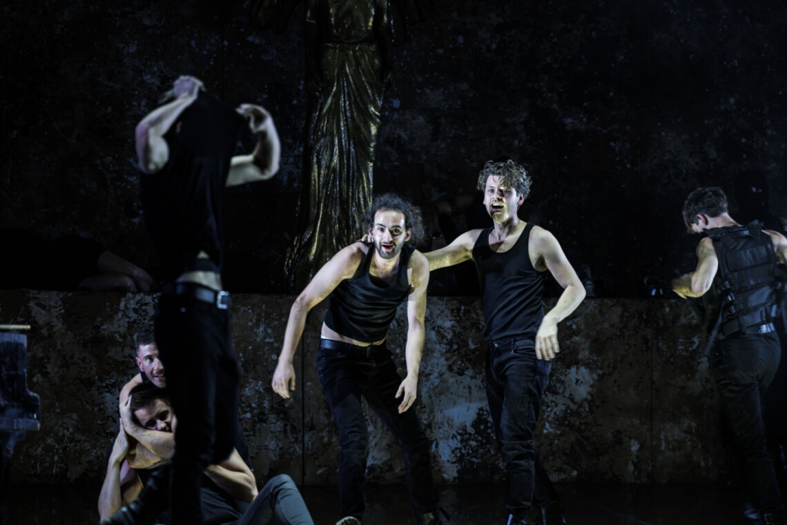 Mężczyźni ubrani na czarno, w centrum dwóch z nich, na pierwszym planie mężczyzna zdejmuje koszulkę