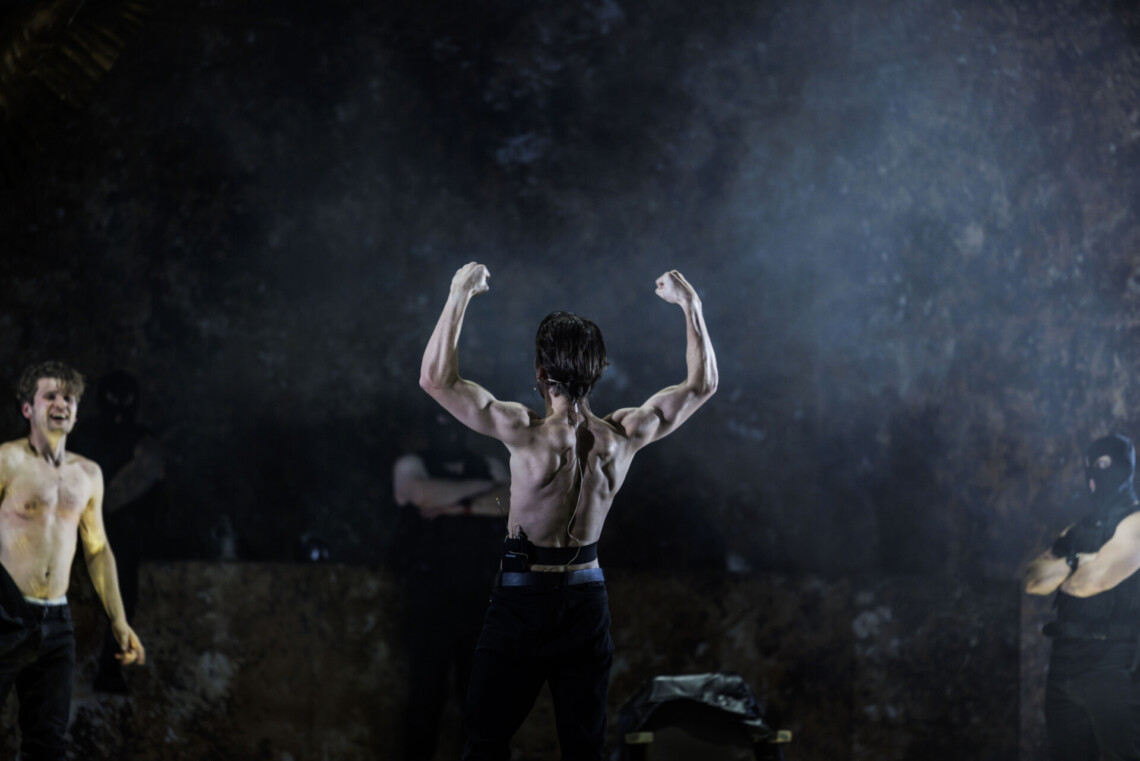 Mężczyzna bez koszulki stojący tyłem, ma uniesione w górę ręce i napina mięśnie