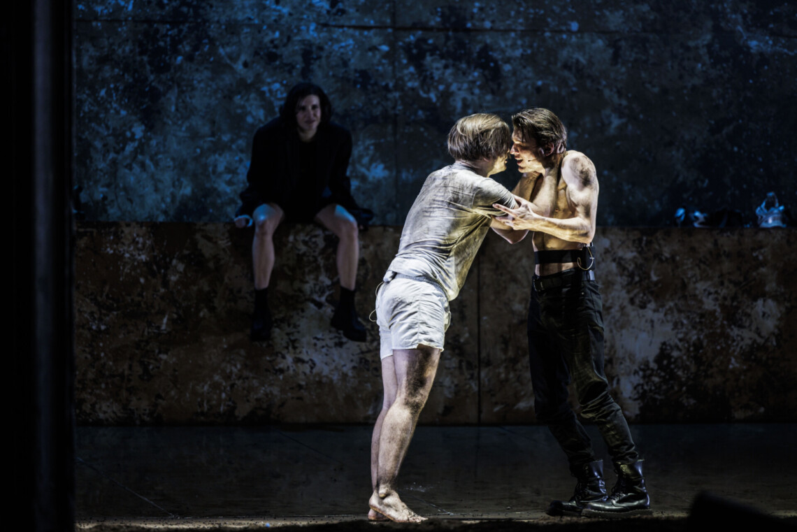 Dwóch mężczyzn stoi naprzeciwko siebie, z lewej ubrudzony mężczyzna ubrany na biało trzyma swoje ręce na szyi i głowie drugiego, drugi mężczyzna jest bez koszulki i opiera swoje ręce na ramionach pierwszego, na drugim planie na podwyższeniu siedzi kobieta ubrana na czarno