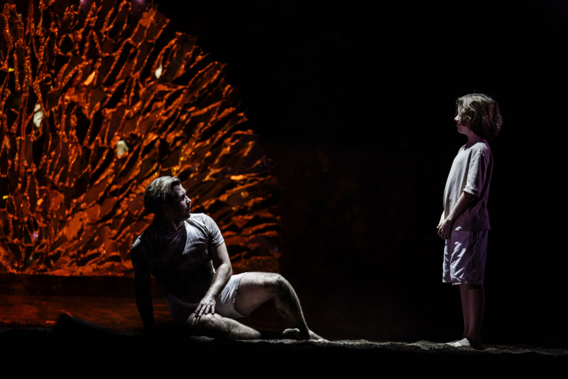 Na podłodze w ziemi siedzi mężczyzna, naprzeciwko niego stoi chłopak, obaj ubrani na biało w krótkie spodenki, za nimi złoty, półokrągły element dekoracji