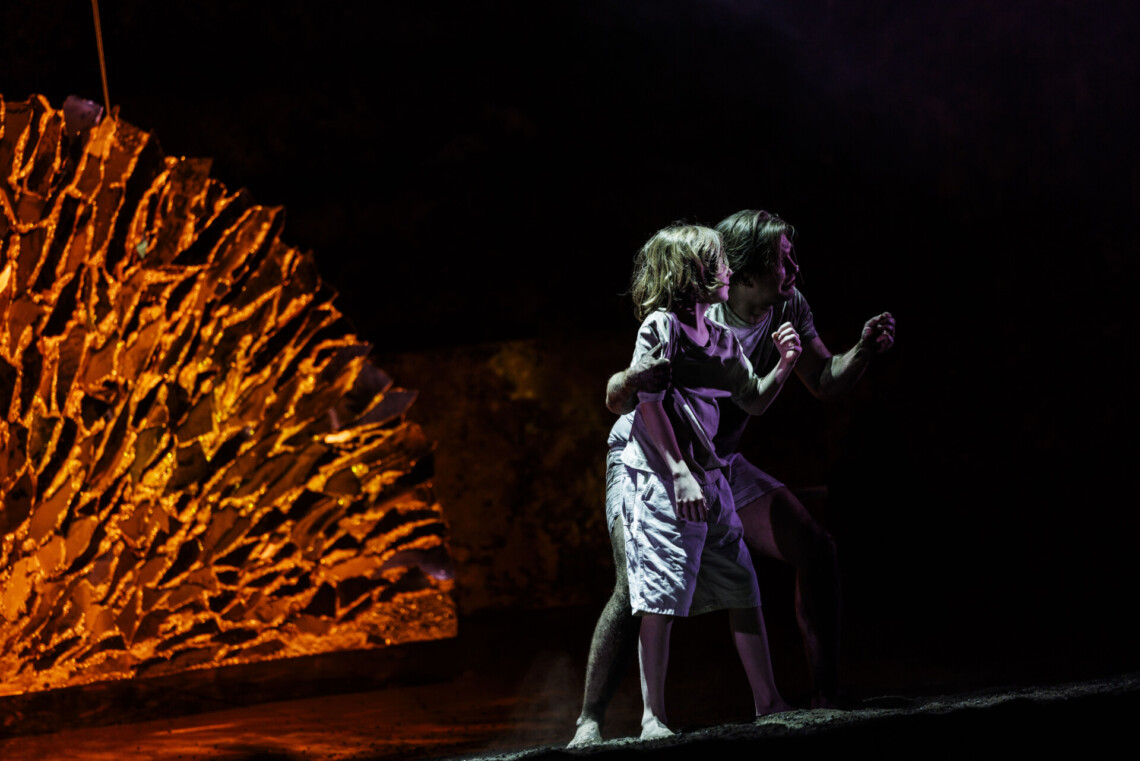 Chłopak i mężczyzna ubrani na biało stoją razem i patrzą w tym samym kierunku, mężczyzna obejmuje chłopaka, za nimi złoty, półokrągły element dekoracji