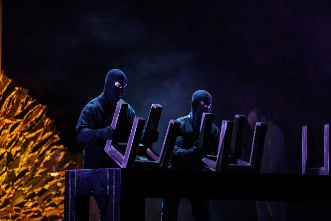 Postacie ubrane na czarno w kominiarkach stoją przy stole, na który kładą krzesła