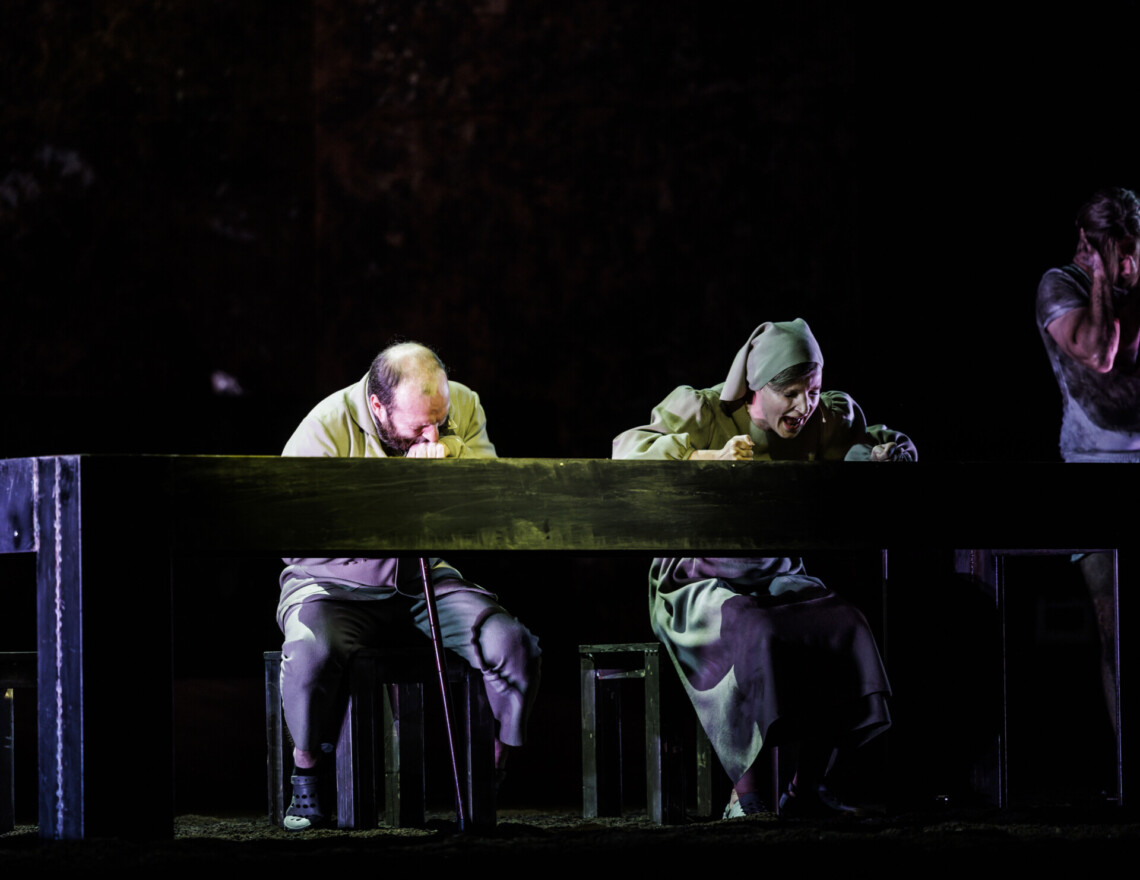 Za długim, czarnym stołem siedzą mężczyzna i kobieta w chuście, mężczyzna ma pochyloną głowę bezpośrednio nad blatem stołu, kobieta ma otwarte szeroko usta jak w krzyku, obok stołu stoi ubrudzony mężczyzna i zatyka uszy