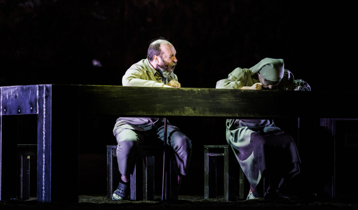 Za długim, czarnym stołem siedzą mężczyzna i kobieta w chuście, mężczyzna ma otwarte szeroko usta jak w krzyku, kobieta ma pochyloną głowę bezpośrednio nad blatem stołu