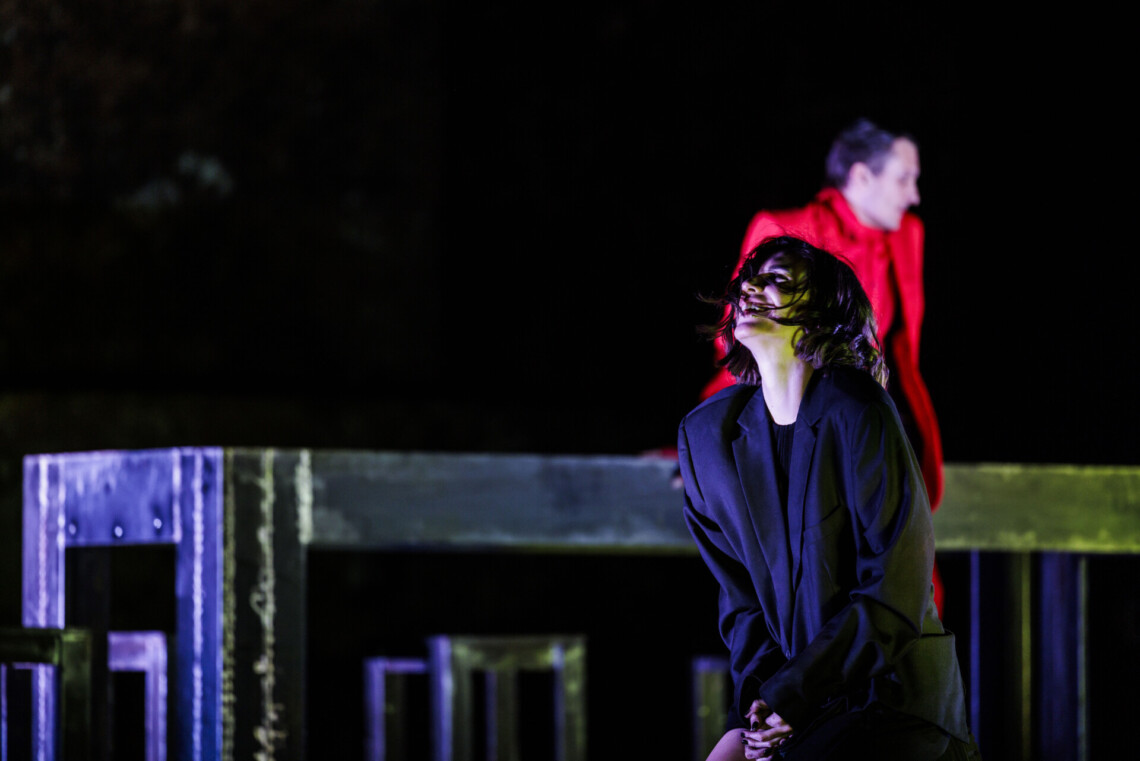 Na pierwszym planie klęczy kobieta ubrana w czarną marynarkę, na drugim planie przy długim, czarnym stole stoi mężczyzna w czerwonym garniturze