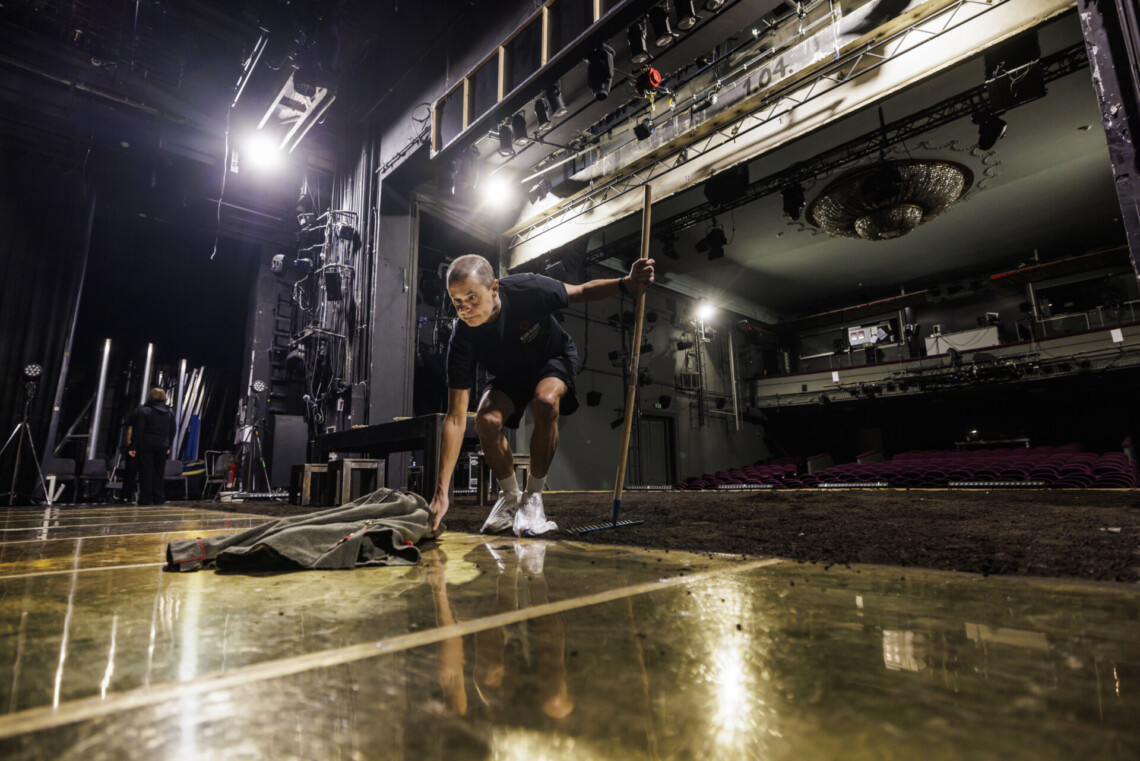 Mężczyzna na scenie podnoszący z podłogi kurtkę, w lewej dłoni trzyma grabki, na części sceny jest pas ziemi, w tle pusta widownia