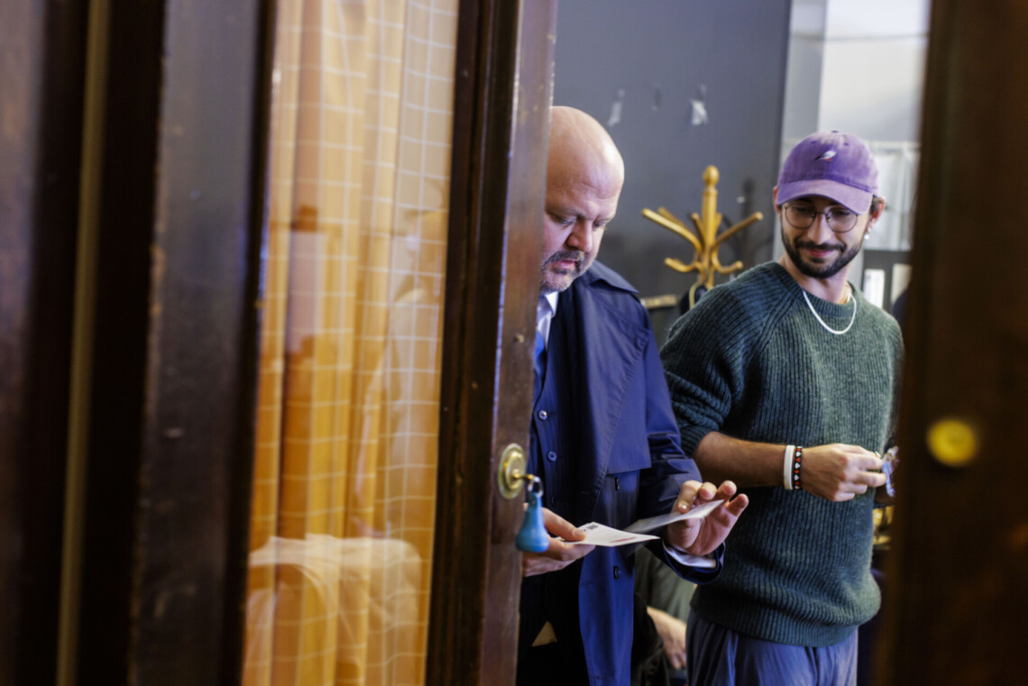 Na pierwszym planie drzwi za nimi, dwóch mężczyzn, pierwszy w płaszczu i garniturze, drugi w swetrze i czapce z daszkiem, elegancki mężczyzna trzyma w dłoniach dwa bilety, drugi patrzy na nie