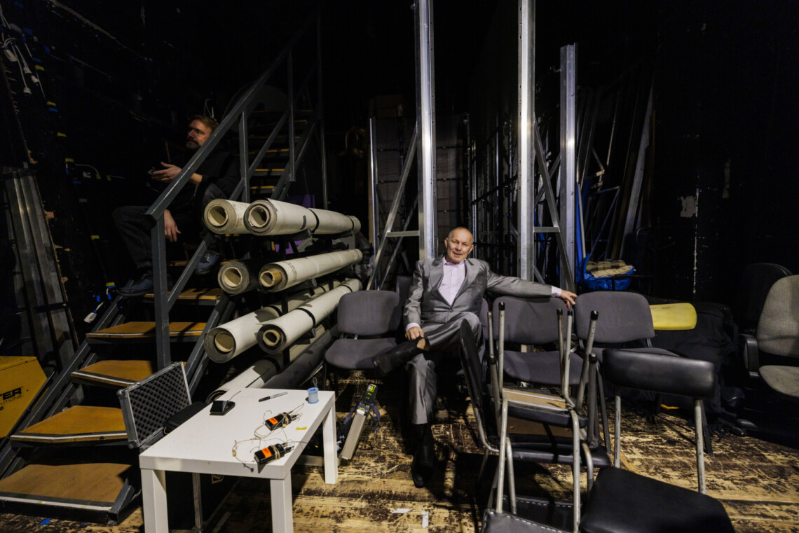 Aktor w garniturze siedzący za kulisami na krześle, wokół niego krzesła, część jest ustawiona jedno na drugie, z lewej strony schody, na których w cieniu siedzi mężczyzna