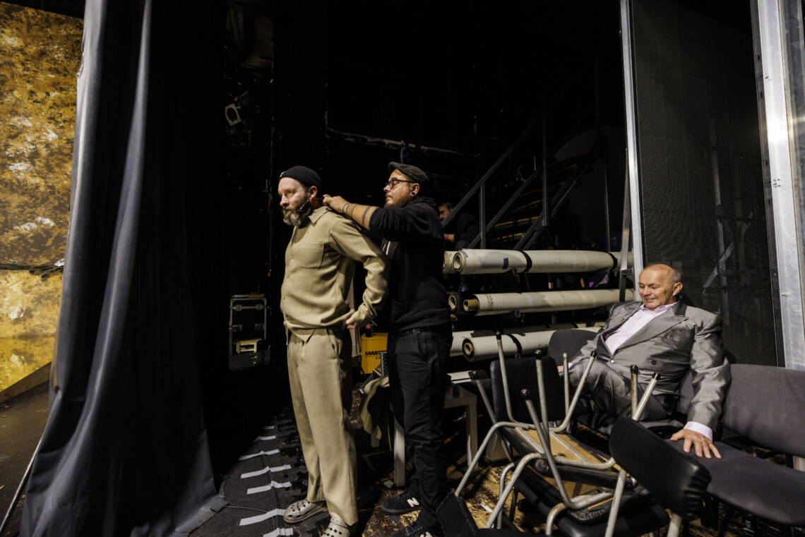 Trzech mężczyzn za kulisami, aktor i pracownik obsługi technicznej stoją jeden z drugim, pracownik zakłada mu mikrofon przy szyi, za nimi na krześle siedzi aktor w garniturze, wokół niego ustawione krzesła