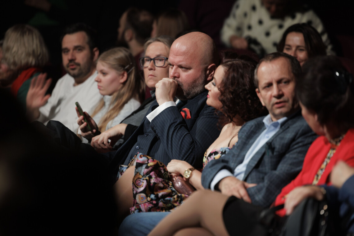 Osoby siedzące na widowni, w centrum elegancko ubrani kobieta i mężczyzna