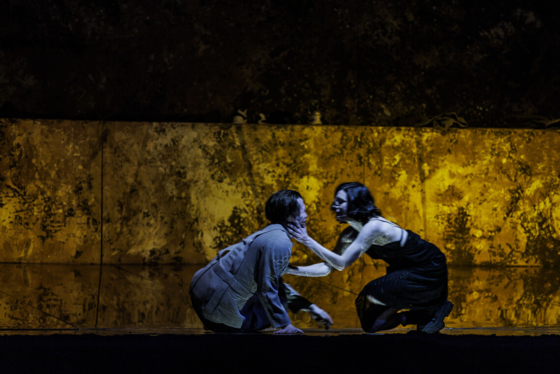 Na podłodze siedzi mężczyzna, na przeciwko niego kuca kobieta, opiera dłoń na jego szyi, a drugą klatce piersiowej, za nimi złota ściana