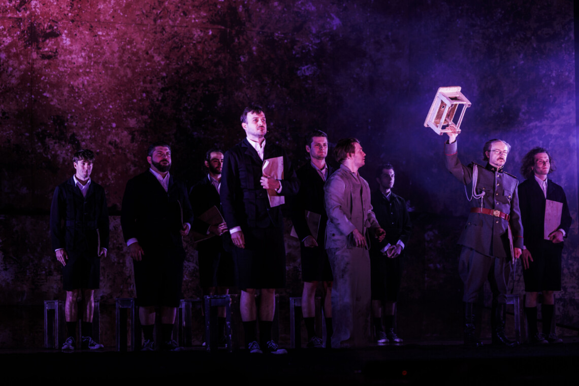 Z lewej strony grupa chłopaków w mundurkach z krótkimi spodenkami, na przodzie jeden ubrany inaczej, z prawej strony mężczyzna w mundurze w prawej ręce trzyma niski, prostokątny taboret, z drugiej jego strony stoi chłopak w krótkich spodenkach