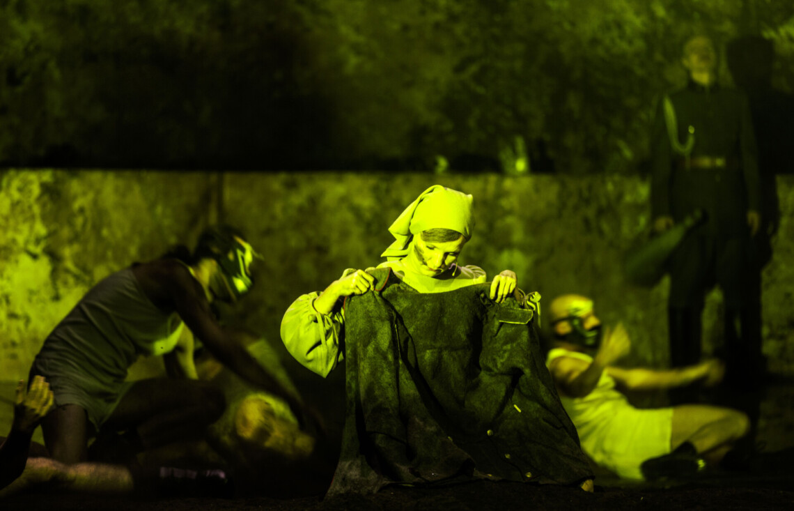 Kobieta w chuście na głowie siedzi na podłodze, przed sobą trzyma kurtkę wojskową na którą patrzy, scena oświetlona na żółto