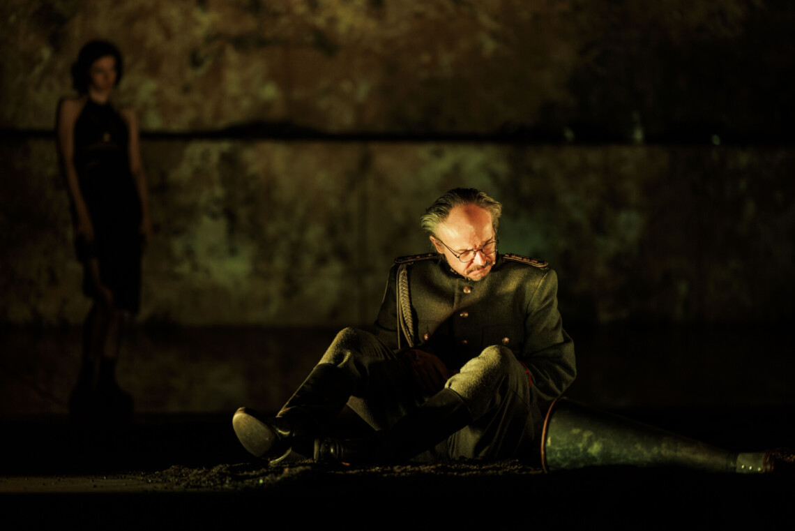 Mężczyzna w mundurze siedzący na ziemi, głowę ma skierowaną w dół, obok niego leży megafon tubowy