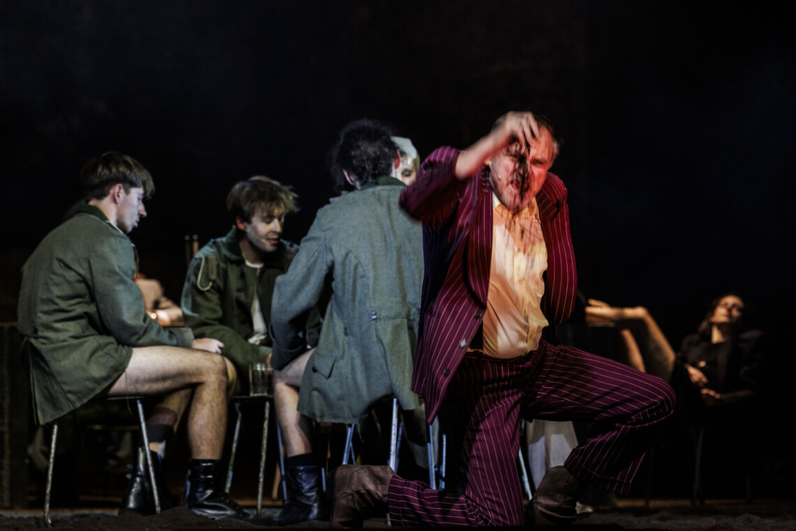 Na pierwszym planie na jednym kolanie klęczy mężczyzna w bordowym garniturze, z prawej dłoni wypuszcza ziemię, na drugim planie na taboretach siedzą mężczyźni w wojskowych kurtkach