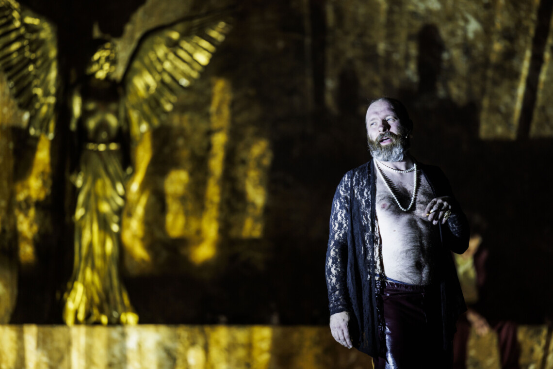 Z prawej strony mężczyzna bez koszulki w zarzuconym na górę koronkowym, rozpinanym swetrze, patrzy w bok, w tle posąg złotego anioła