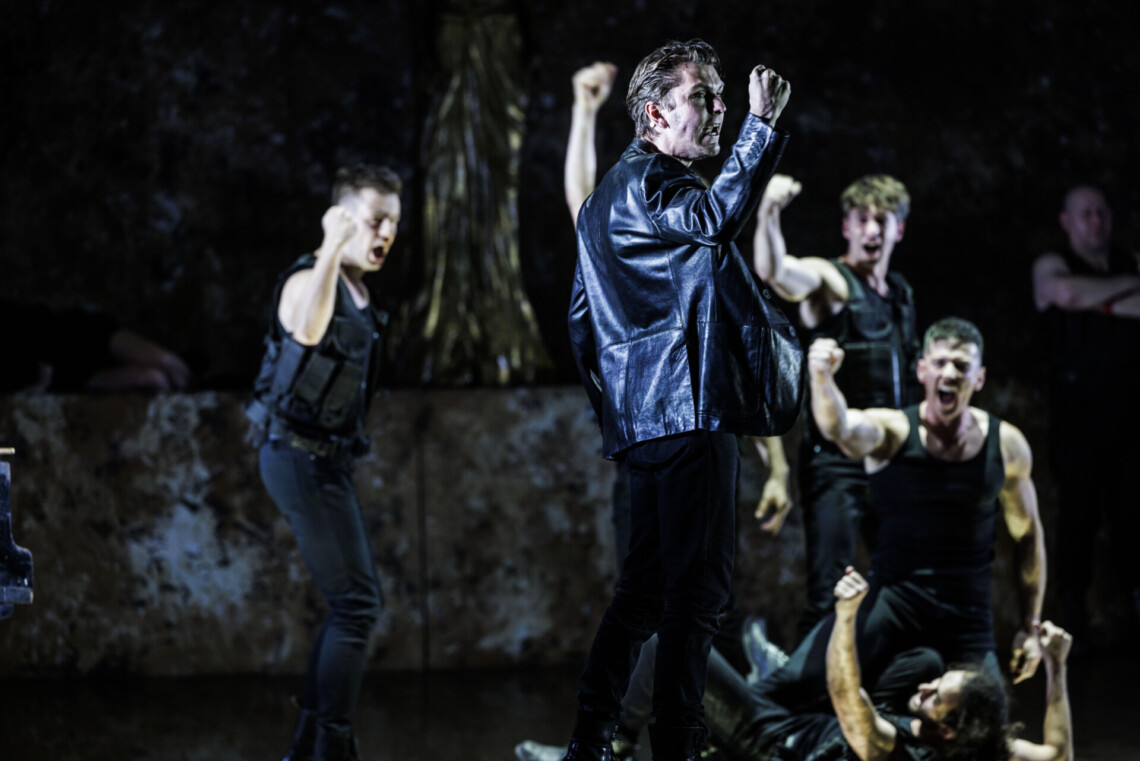 Na pierwszym planie mężczyzna stojący bokiem ze zgiętą ręką w łokciu, uniesioną w górę i zaciśniętą pięścią, na drugim planie czterech mężczyzn w podobnej pozie, przy czym jeden leży na podłodze, a drugi na nim siedzi, tło ciemne