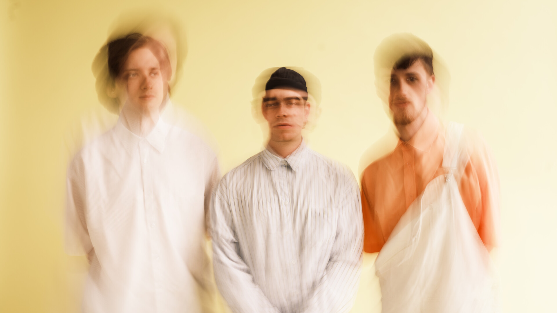 Trzech mężczyzn ubranych na biało, rozmazani, w środku najniższy mężczyzna, za nimi jasne tło