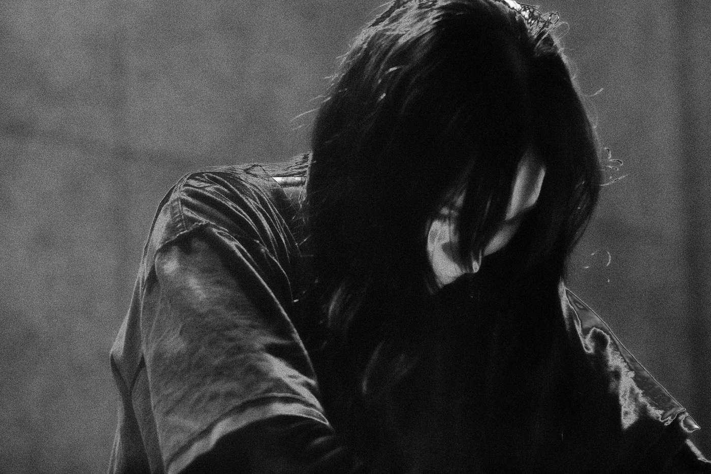 Osoba z ciemnymi włosami ze spuszczoną głową, włosy przykrywają twarz, z tyłu szare tło, zdjęcie w odcieniach szarości i czerni